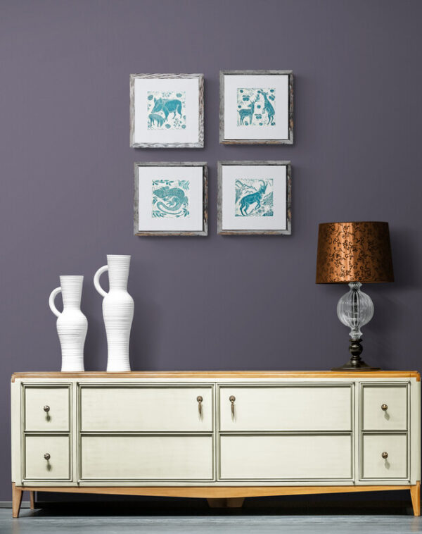 Four teal wildlife lino prints on a lilac wall above a contemporary low cupboard, beteen two white vases and a glass lamp with a bronze metal shade.